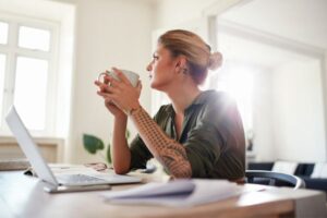 Decisões estratégicas. Mulher loura sentada em frente a um laptop, segurando uma xícada branca.