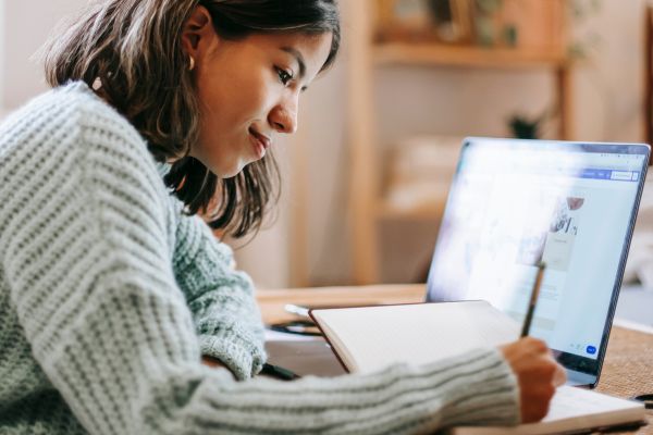 Mulher sentada em frente ao laptop, escrevendo em um caderno. Foco e concentração.